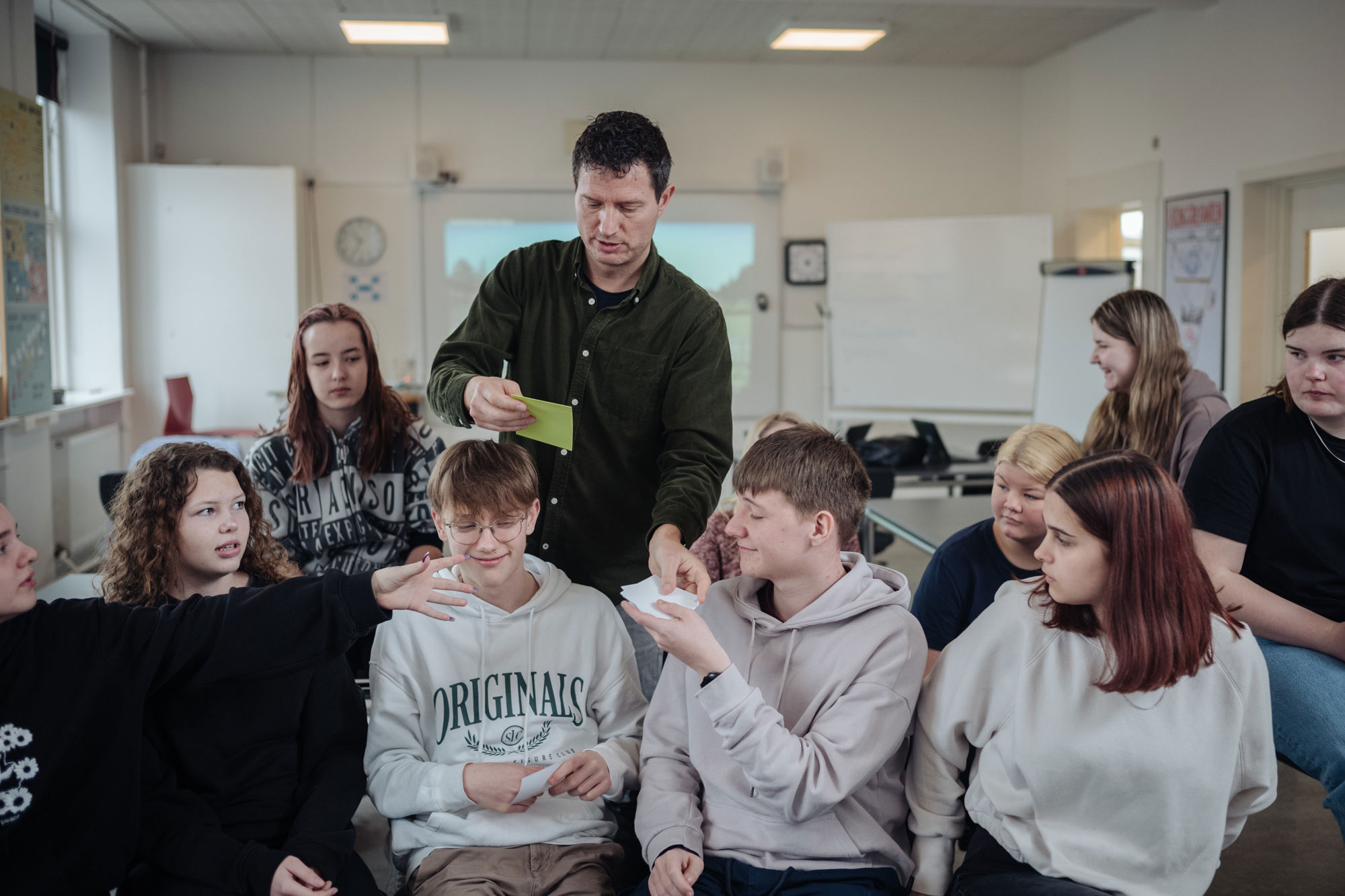 Billede af undervisningsituation med skolelederen og en gruppe elever, der modtager papirlapper.